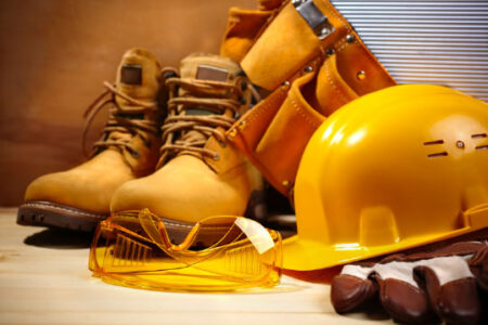 Close-up view of construction personal protective equipment, including safety goggles, a hard hat, a leather toolbelt, and steel-toe boots.