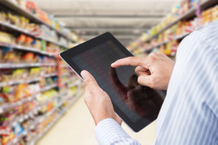 Over-the-shoulder view of a person holding a tablet, reviewing inventory in a grocery store aisle.