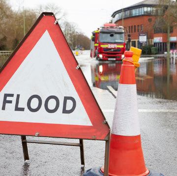 A “flood” warning sign sits on a flooded city street, with a business building and fire truck in the background