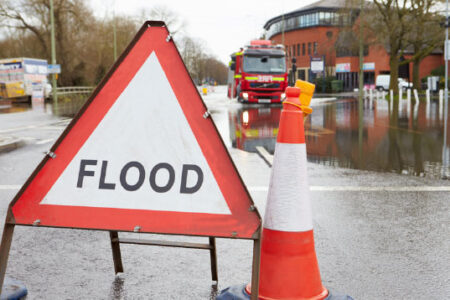 A “flood” warning sign sits on a flooded city street, with a business building and fire truck in the background